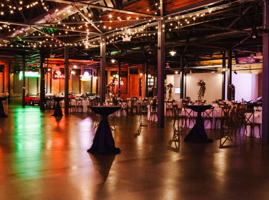 A large rental venue with classic cars, tables high and low, and hanging lights. 
